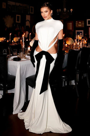 Woman in a white evening gown with a large black bow standing in a dimly lit room with tables and candles.